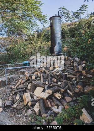 Haufen gehackten Brennholzes, draußen neben einem Metallholzofen in einem grünen ländlichen Hof mit Bäumen und Büschen gestapelt Stockfoto