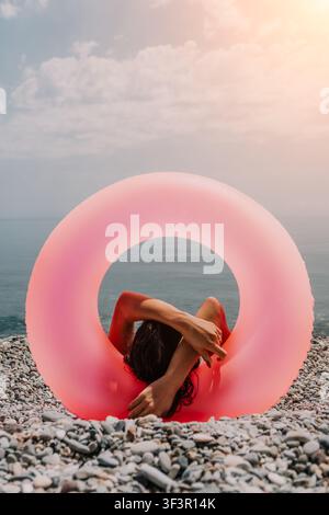 Schwimmring-Strandentspannung, Frau sitzt auf felsiger Küste mit rosa aufblasbarem Ring, der das Gesicht vor der Sonne schützt, Sommerurlaub-Konzept Stockfoto