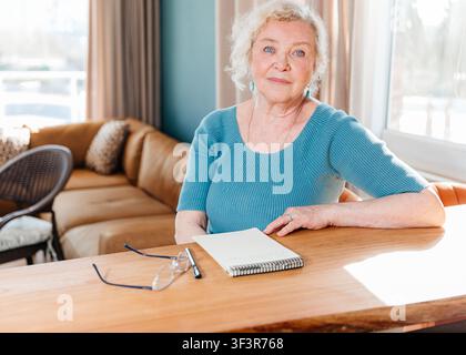 Porträt einer selbstbewussten Seniorin, die an einem Tisch mit einem Notizbuch und einer Brille in einem hellen Wohnrauminneren sitzt. Stockfoto