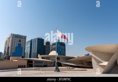 Doha, Katar - 14. Februar 2026: Die qatarische Nationalflagge weht vor dem Nationalmuseum von Katar, mit modernen Gebäuden von Al Salata. Stockfoto