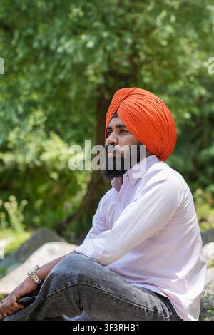 Vertikale Seitenansicht eines jungen Sikh-Mannes mit einem orangefarbenen Turban, der mit einem schockierten und überraschten Gesichtsausdruck sitzt, vor einem unscharfen Hintergrund. Stockfoto