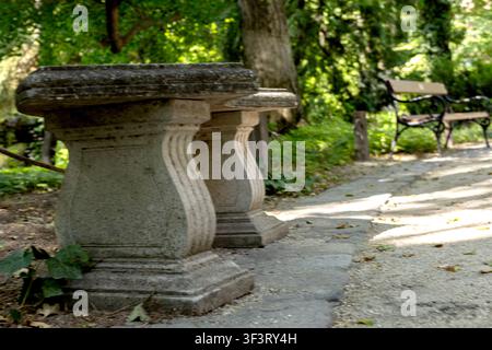 Eine Steinbank steht neben einem schattigen Gehweg in einem grünen Park. Sonnenlicht dringt durch die Bäume und fällt auf den Kies und die Pflastersteine. Stockfoto