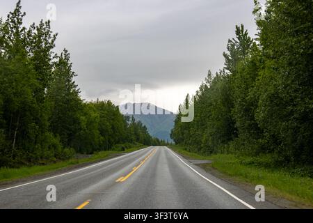 Pittoreske Bergstraße, die sich durch grüne Bäume zu einem fernen Gipfel unter bewölktem Himmel im Hatcher Pass, Alaska, USA erstreckt Stockfoto