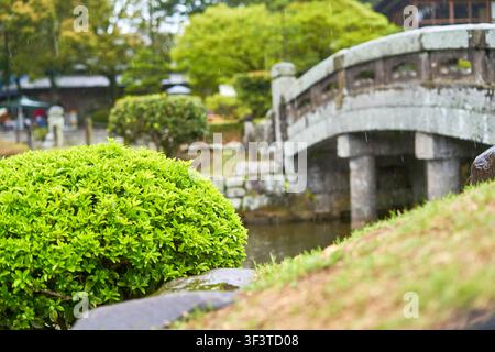 Eine Nahaufnahme von lebendigem grünem Buschlaub im scharfen Fokus im Vordergrund, mit einer traditionellen japanischen Steinbrücke und hölzernen Geländern im Hintergrund Stockfoto