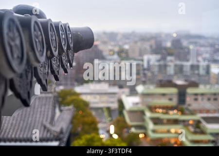 Eine Reihe schwarzer, münzbetriebenen Fernglasstationen, die auf einem Metallgeländer montiert sind, mit Regentropfen bedeckt, mit Blick auf eine verschwommene urbane Stadtlandschaft an einem bewölkten Tag Stockfoto