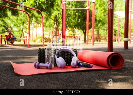 Outdoor-Fitnessgeräte mit einer roten Yogamatte, Hanteln, Wasserflasche und Kopfhörern auf einer dunklen Oberfläche Stockfoto