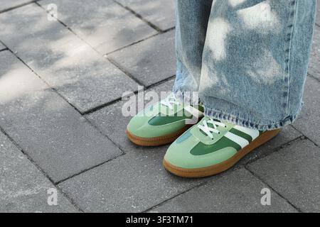 Person, die helle Denimjeans und grüne Turnschuhe mit weißen Streifen trägt, steht auf grauem Pflaster im Freien, die Person steht auf einem grauen Pflaster Stockfoto