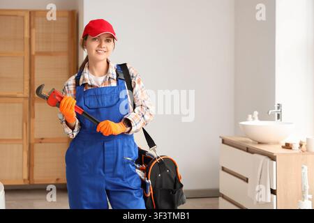 Lächelnde junge Klempnerin in blauen Latzhosen und roter Mütze, die einen Rohrzang hält, in einem modernen Badezimmer. Sie steht selbstbewusst in einem modernen Badezimmer Stockfoto