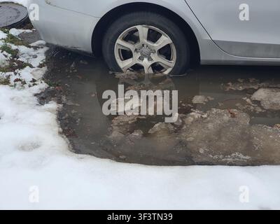 Ein silberner Audi mit einem teilweise in einem großen, mit Wasser gefüllten Schlagloch versunkenen Hinterrad auf einer beschädigten Asphaltstraße. Schmelzender Schnee und Eis umgeben Stockfoto