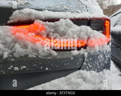 Nahaufnahme der Rückseite eines dunkel gefärbten SUV oder Crossover-Fahrzeugs, das mit dickem, frischem Weißschnee bedeckt ist. Das moderne LED-Rücklicht ist beleuchtet, Stockfoto