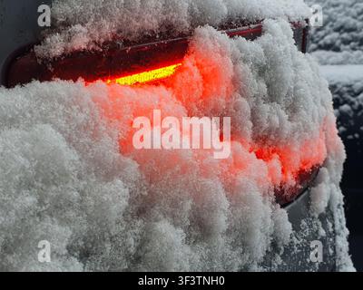 Nahaufnahme eines Hecklichts eines Autos, das teilweise mit dickem, frischem Schnee bedeckt ist. Das rote Bremslicht leuchtet und erzeugt ein orange-rotes Glühen Stockfoto