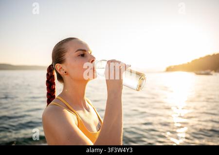 Seitenansicht einer sportlichen jungen Frau, die am Meer bei Sonnenaufgang aus einer Glasflasche trinkt und Outdoor-Fitness, Hydrierung und ein gesundes Sommerleben genießt Stockfoto