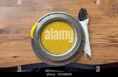 Eine warme, tröstliche gelbe Linsensuppe, serviert in einer dekorativen Metallschale mit aufwendigen Mustern, begleitet von einer Zitronenscheibe und Besteck auf Holz Stockfoto