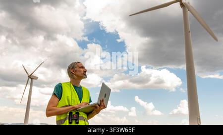 Malerneuerungsingenieur und Feldtechniker mit Sicherheitsweste überprüft die Leistung einer Windturbine mit einem Laptop während der Wartung an einem windigen Kraftwerksstandort Stockfoto
