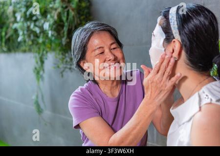 Lächelnde ältere Frau hilft einer jungen Frau sanft dabei, eine Gesichtsmaske zu Hause aufzutragen, zeigt Hautpflegeroutine, familiäre Bindung, Selbstpflege und Wellness Stockfoto