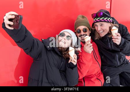 Drei Freunde in Winterjacken machen ein fröhliches Smartphone-Selfie vor einer leuchtend roten Wand, lachen zusammen, während sie Eisbecher halten, während einer Stockfoto