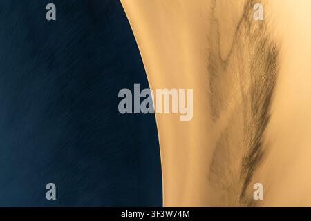 Minimaler abstrakter Naturhintergrund mit weiten Dünenmustern und feinen Sandtexturen, bei dem sanfte goldene Kurven auf tiefe blaue Schatten treffen, für Ruhe Stockfoto