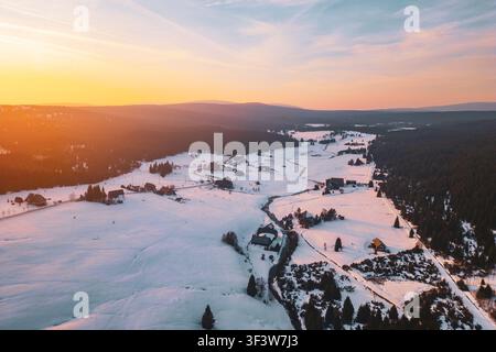 Luftaufnahme zeigt das Bergdorf Jizerka im Winter. Schnee bedeckt die Landschaft, und Häuser sind über das Tal verstreut, während die Sonne im Hintergrund untergeht. Stockfoto