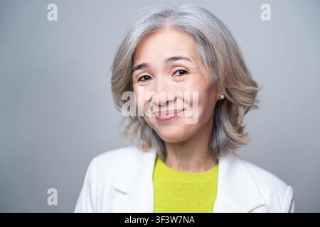 Nahaufnahmeporträt einer lächelnden älteren asiatischen Frau mit grauen Haaren in einem weißen Blazer und hellem Oberteil, die selbstbewusst in weichem Studiolicht posiert, vor Stockfoto