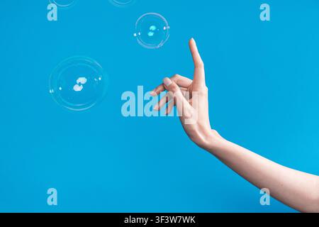 Nahaufnahme von kaukasischen Frauenhänden, die nach schwebenden Seifenblasen auf hellem blauem Hintergrund greifen und darauf zeigen, minimalistisches Studio-Konzept mit Kopierplatz Stockfoto