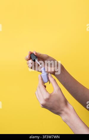 Nahaufnahme von kaukasischen Frauenhänden, die eine offene Flasche Lavendel-Nagellack mit Pinsel-Applikator halten, isoliert auf einem hellgelben Hintergrund mit Kopie Stockfoto