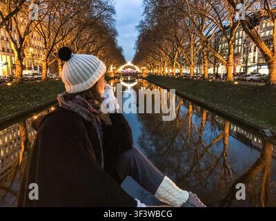 Eine elegante Frau in Winterkleidung sitzt am Kanal an der Königsallee, umgeben von beleuchteten Bäumen und festlichen Weihnachtslichtern Stockfoto