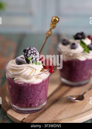 Vertikale Food-Fotografie eines geschichteten Beeren-Smoothie-Desserts, gekrönt mit Schlagsahne, Brombeere und roten Johannisbeeren auf einem rustikalen Holztisch Stockfoto