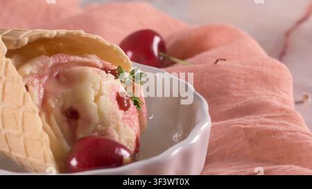 Cremiges Eis in einer knusprigen Waffeltüte ruht in einer weißen Schüssel, garniert mit frischen Kirschen und einem Thymianzweig unter sanftem natürlichem Licht. Stockfoto