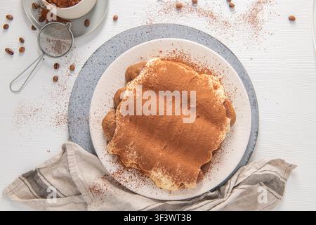 Eine Ansicht von oben auf ein köstliches hausgemachtes Tiramisu, das mit Kakaopulver bestäubt ist, umgeben von Kaffeebohnen, einem kleinen Sieb und einer Tasse, alles arrangiert auf einem Wh Stockfoto