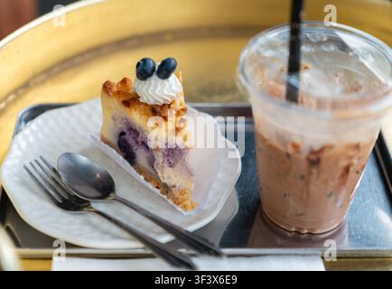 Hohe Winkelaufnahme eines Backwarendesserts mit einem Stück Blaubeerkuchen und einem zum Mitnehmen bestimmten Becher mit Eismilchtee oder Kaffee auf einem Messingtisch. Stockfoto