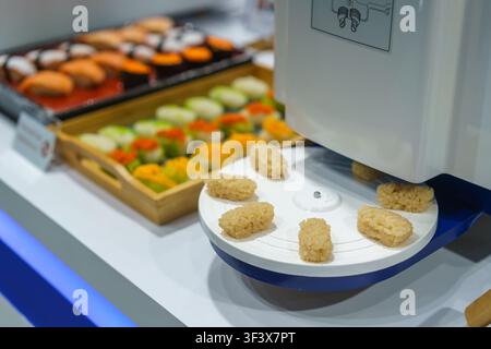 Nahaufnahme einer automatisierten kommerziellen Sushi-Maschine, die perfekt geformte Nigiri-Reiskugeln auf ein rotierendes Tablett abgibt, mit fertigem Sushi im Hintergrund Stockfoto