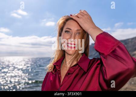 Frau Meer lächelt glückliche reife Frau in einem burgunderroten Seidenhemd, die an einem Strand mit funkelndem Ozeanwasser im Hintergrund steht Stockfoto