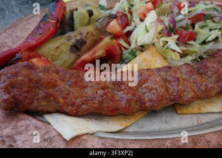 Traditioneller Adana-Kapsalonspieß serviert mit gegrilltem Gemüse und frischem Salat, saftigem Hackfleisch, authentische türkische Küche, geschmackvolles Barbecue-Gericht Stockfoto