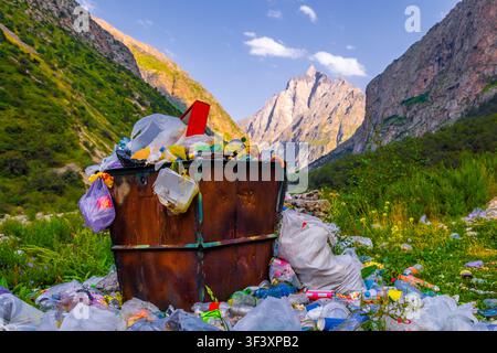 Überquellender Mülleimer gefüllt mit Plastiktüten, Flaschen und Behältern stört die natürliche Schönheit eines Bergtals. Üppiges grünes Laub und br Stockfoto