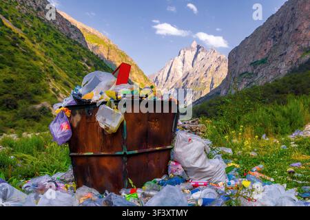 Überquellender Mülleimer gefüllt mit Plastiktüten, Flaschen und Behältern stört die natürliche Schönheit eines Bergtals. Üppiges grünes Laub und br Stockfoto