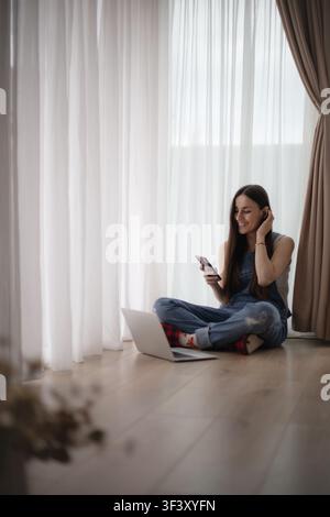 Lächelnde junge Frau berührt einen kabellosen Ohrhörer, während sie ein Smartphone neben einem offenen Laptop zu Hause benutzt, mit einem weißen Hund im Hintergrund, der unscharf ist. Stockfoto