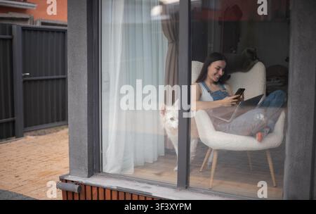 Blick von außen durch ein großes Fenster einer lächelnden jungen Frau, die ein Smartphone neben einem Laptop benutzt, weißer Hund und graue Katze in einem modernen Zuhause. Stockfoto