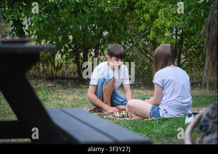 Zwei Teenager spielen Schach, während sie auf einer Picknickdecke im Garten entspannen. Konzept von Freundschaft, Familienfreizeit, Bildung, Strategie und Gesundheit Stockfoto