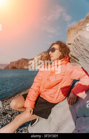 Frau Strand Sonnenuntergang junge Frau in orangefarbener Pufferjacke sitzt an einem felsigen Strand und genießt den Ausblick zur goldenen Stunde am Meer Stockfoto