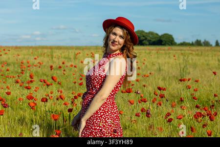 Schöne rothaarige Frau lächelt über die Schulter, während sie in einem blühenden Mohn- und Gerstenfeld posiert, in einem roten Kleid und passendem Hut. Stockfoto