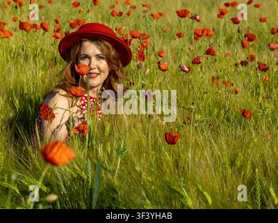 Porträt einer fröhlichen rothaarigen Frau, die einen roten Hut und ein rotes Kleid trägt und unter hellen Mohnblumen und üppigem grünem Gerste in einem Sommerfeld sitzt. Stockfoto