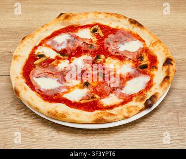 Ganze Prosciutto-Pilz-Pizza mit geschmolzenem Mozzarellakäse und Tomatensoße auf weißem Teller auf Holztablett. Stockfoto