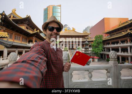 Zufriedener Tourist macht ein lustiges Selfie mit der chinesischen Flagge an der berühmten Touristenattraktion in China. Asiatische Sommerreise. Junger Mann macht ein Handyfoto Stockfoto