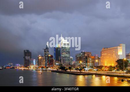 Weite nächtliche Stadtlandschaft mit beleuchteten modernen Türmen, Ufergebäuden, ruhigen Spiegelungen auf dem Wasser und dramatischen Sturmwolken über den geschäftigen Innenstadtlichtern entlang der Uferpromenade, mit Skyline, Stadt Stockfoto