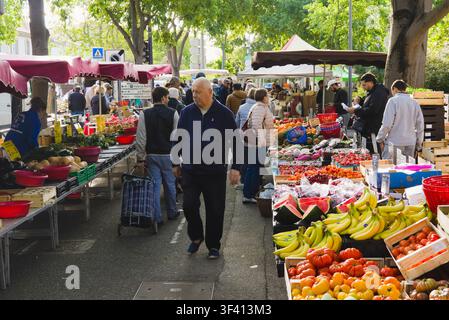 Menschen durchstöbern Obst- und Gemüsestände auf einem geschäftigen Freiluftmarkt unter grünen Bäumen in Arles, Frankreich. Stockfoto