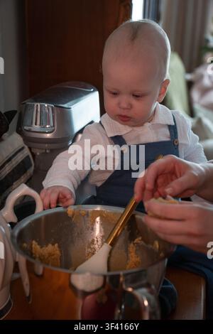 Neugieriges Baby probiert hausgemachten Mandelkuchenteig, während die Familie zusammen in der gemütlichen Küche zu Hause Dessert zubereitet. Kindheit, Familienbacken, Elternschaft, Stockfoto