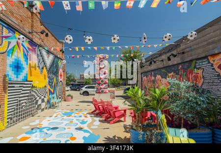 Eine lebendige Gasse zeigt Ziegelmauern, die mit farbenfrohen Wandgemälden bedeckt sind, rote Stühle und hängende Fußballball-Dekorationen unter einem klaren Himmel. Stockfoto