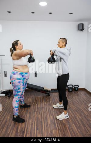 Übergewichtige Frau hebt Kettlebells mit Personal Trainer im Fitnessstudio Stockfoto