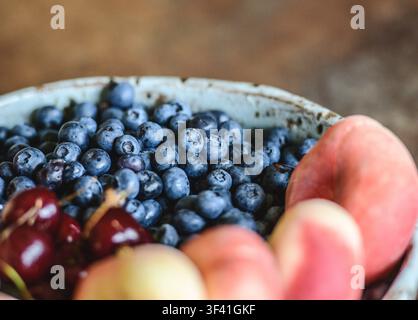 Keramikschale auf einem Holztisch gefüllt mit frischen Blaubeeren neben Kirschen und Pfirsichen Stockfoto