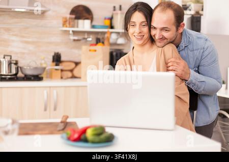Romantisches Paar verwendet Laptop in der Küche, um nach einem Frühstücksrezept zu suchen, zu Hause mit moderner WLAN-Drahtlos-Internet-Technologie, Laptop-Arbeit, Computerarbeit und Fernarbeit verbinden. Stockfoto